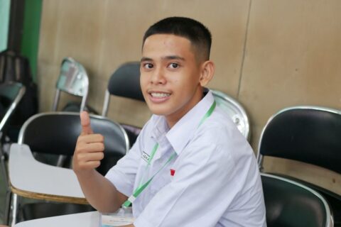 a young man sitting at a table giving a thumbs up